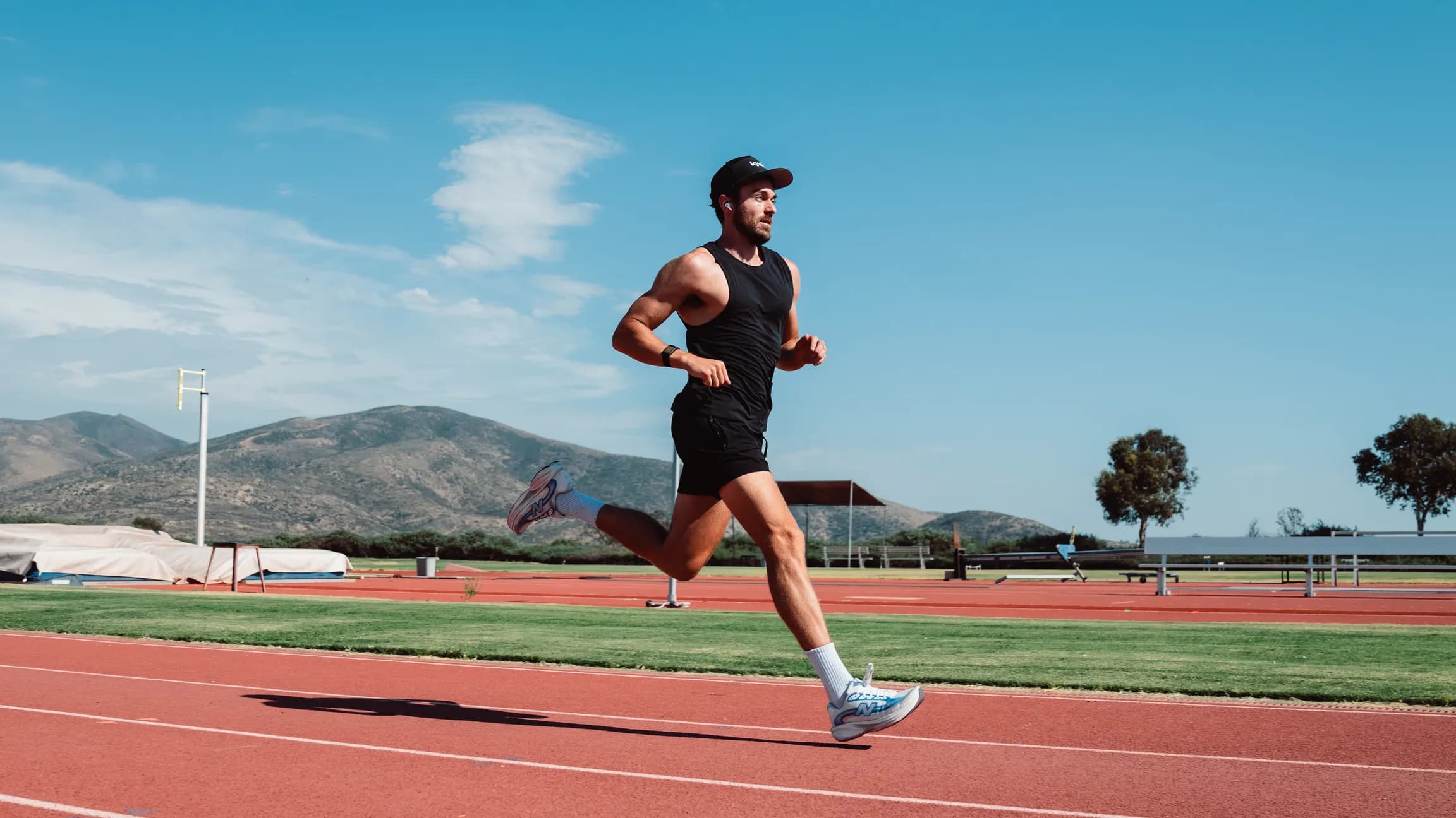 Runner mid-stride on the track in a Kyde cap, mountains and blue sky behind